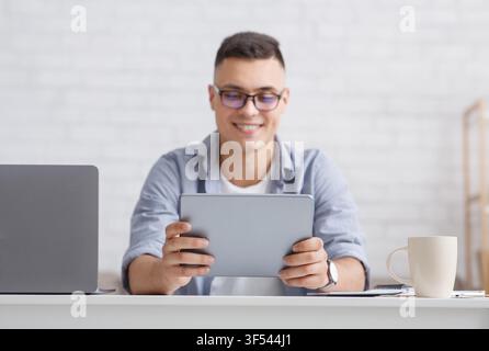 Man looking at web camera making conference video call. Smiling teacher holds digital tablet, sitting at table with laptop in living room interior Stock Photo