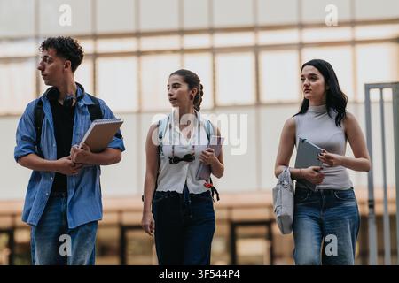 Three students walking on campus carrying notebooks and backpacks while talking between classes Stock Photo