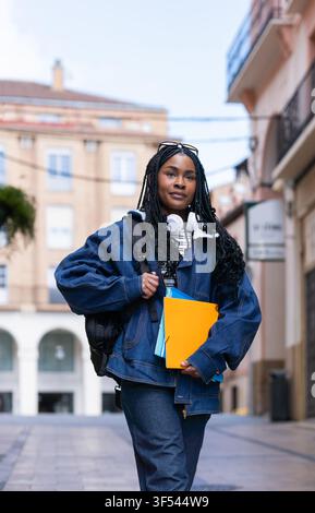 Confident young woman student walking down the street, carrying folders and backpack Stock Photo