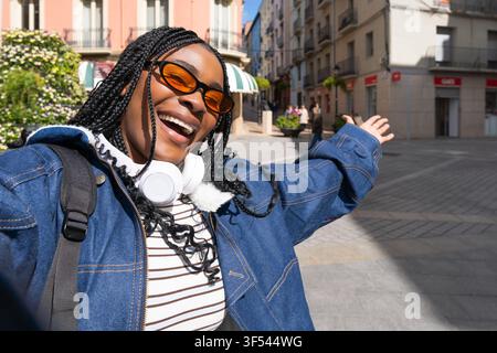 Young woman with braids, sunglasses and headphones taking a selfie with arms outstretched, enjoying her trip in a sunny city during spring Stock Photo
