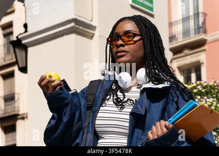 Young black student with headphones and sunglasses holding books and an apple, enjoying a sunny day in the city Stock Photo