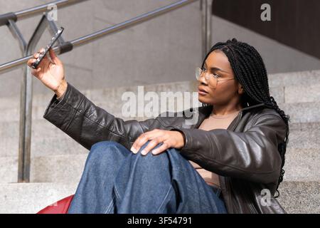 Stylish young woman taking a selfie with her smartphone while sitting on steps in an urban setting Stock Photo