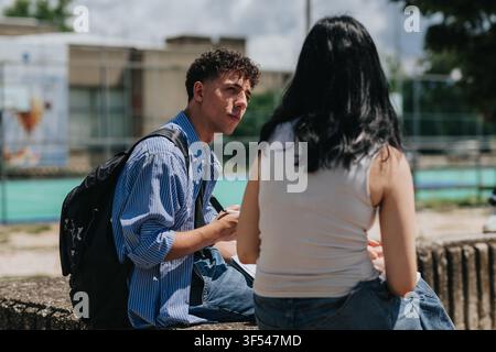 Two students talking outdoors on a campus bench with backpacks and notebooks Stock Photo