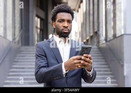 Young african ethnicity businessman in a formal suit using his mobile phone, standing in front of modern office building stairs, representing communic Stock Photo
