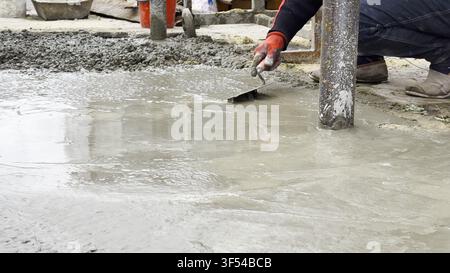 Male hands in protective gloves uses trowel to spread and level fresh cement in yard. Material is carefully applied to surface as part of preparation Stock Photo