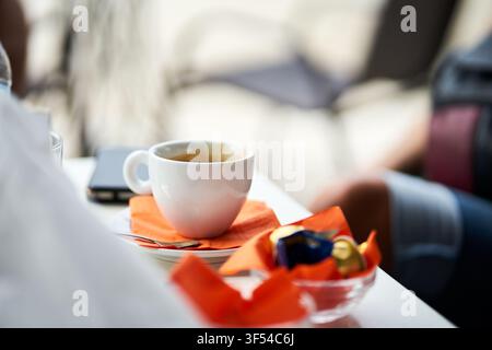A close-up view of a ceramic white coffee cup resting on a saucer with bright orange napkins and chocolates on a cafe table during a break. Stock Photo