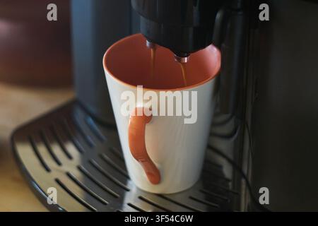 Fresh hot coffee is pouring from the nozzles of an automatic espresso machine directly into a white and orange ceramic mug standing on the drip tray i Stock Photo