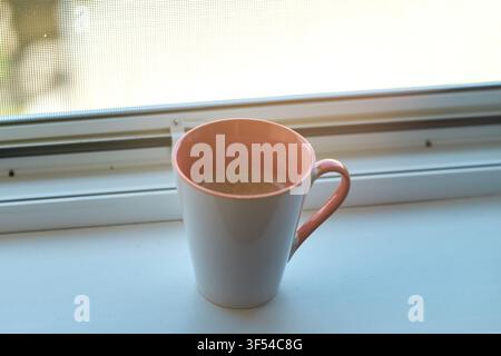 Close-up view of a ceramic coffee mug filled with hot beverage placed on a white windowsill near a window with soft natural light coming through. Stock Photo