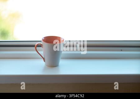 A close-up view of an empty ceramic coffee mug standing on a clean white windowsill with bright natural light coming through the window glass, creatin Stock Photo
