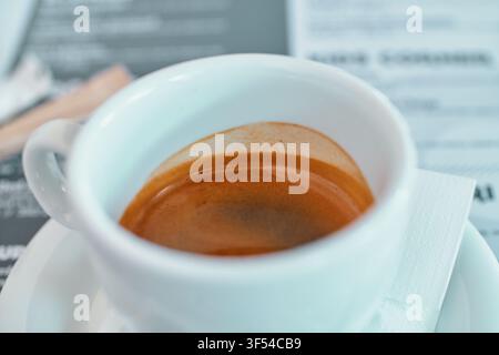 A close-up shot of a small white ceramic cup filled with rich espresso coffee with a thick golden crema, sitting on a matching saucer on a cafe table. Stock Photo