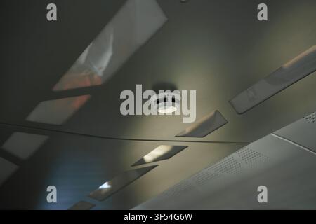 Smoke detector installed on a textured train ceiling with recessed lighting panels, highlighting fire safety and building security systems Stock Photo
