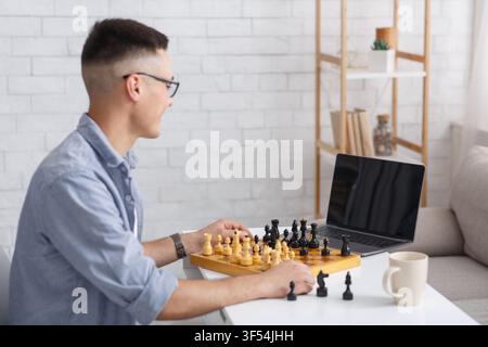 Smart guy and hobby in free time on self-isolation. Happy chess player in glasses with chess board looking at laptop with blank screen in living room Stock Photo