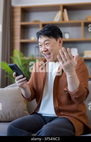 Asian man sitting indoors, checking smartphone, receiving good news, expressing surprise and happiness with an open mouth and raised hand, celebrating Stock Photo