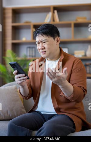 Asian man sitting on a sofa expressing confusion and frustration while using his mobile phone, experiencing problems with communication and digital te Stock Photo