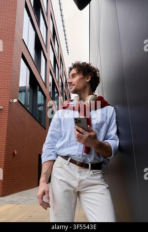 A well dressed man stands confidently against a modern building, checking his phone leisurely. Stock Photo
