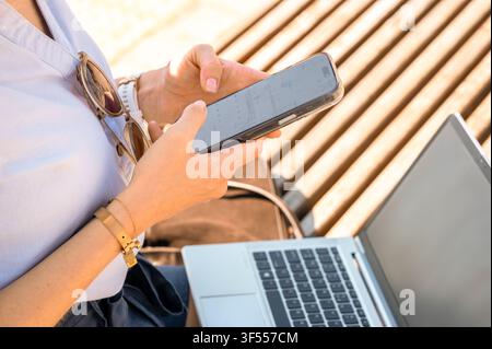Close up of woman hands holding smartphone and checking calendar outdoors. Freelancer working remotely on laptop online Stock Photo
