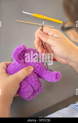 Woman hands sewing eyebrows with black thread and needle on purple plush elephant. Handcrafted amigurumi face details creation Stock Photo