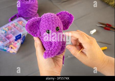 Hands holding purple crocheted plush elephant toy. Handmade amigurumi stuffed animal creation with plush yarn and crafting supplies background Stock Photo