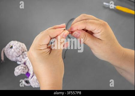 Woman hands threading black thread into needle for sewing handmade toy. Close up of master preparing materials during amigurumi craft lesson Stock Photo
