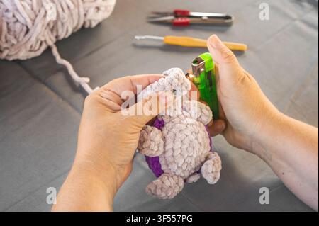 Close up of woman hands using lighter to melt safety eye stem on plush crochet turtle toy. Creative handmade hobby craft and amigurumi making process Stock Photo