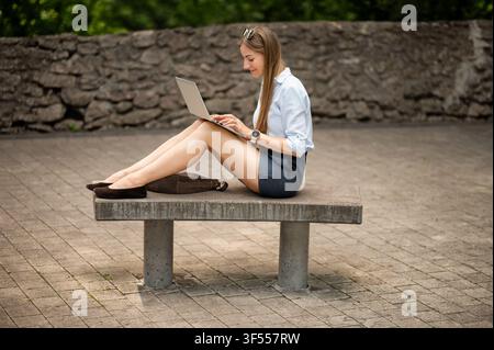 Young female student sitting on concrete bench and typing on laptop outdoors. Remote education and online learning in city park Stock Photo