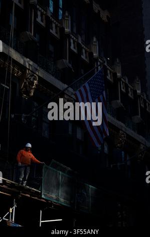 An orange jacket construction worker stands on a scaffold directly underneath a large American flag on a historic NY building Stock Photo