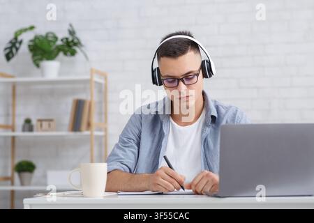 Busy young man with glasses makes notes near laptop in living room interior Stock Photo