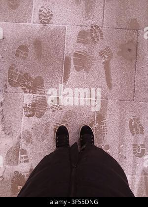 Top down view of black winter boots on a tiled sidewalk covered in melting snow and muddy tracks Stock Photo