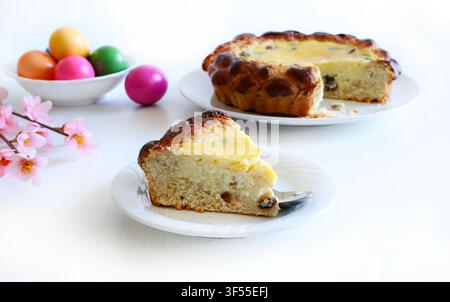Slice of traditional Romanian Easter Pasca cake with cottage cheese and raisins, next to colored eggs on white table. Easter and holiday concept. Stock Photo