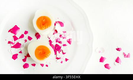 Halved hard boiled egg on white plate with vibrant pink shell pieces on a white background. Easter food concept, overhead view. Stock Photo