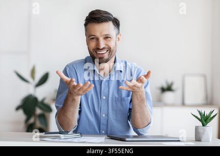 Successful handsome businessman sitting at desk in office, talking at camera Stock Photo