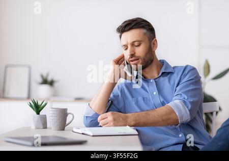 Millennial Male Entrepreneur Talking On Cellphone And Taking Notes In Office Stock Photo