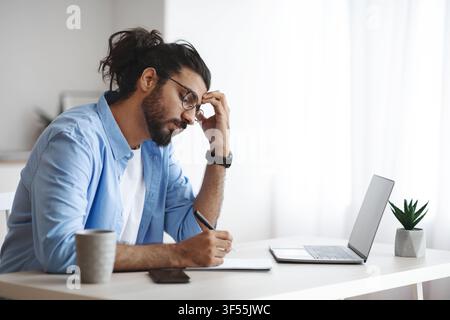 Concentrated Indian Freelancer Guy Noting Ideas To Notepad, Working At Home Office Stock Photo