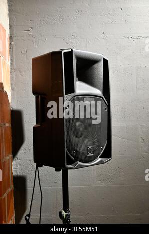 Close-up view of a professional black stage audio speaker mounted on a metal stand against a textured concrete wall in a venue or studio setting.  Stock Photo