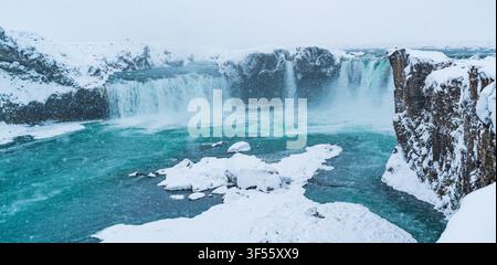 Stunning frozen waterfall flowing into vibrant turquoise river surrounded by snow covered rocky cliffs in winter Stock Photo