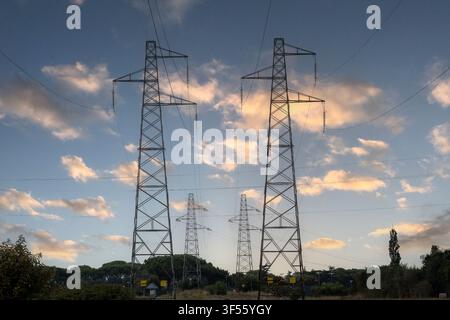 Low angle view of high voltage power pylons and electrical lines against a blue sky with golden clouds at sunset. Stock Photo