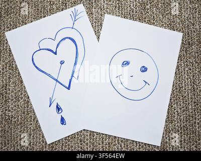 Two sheets of white paper show simple drawings. One sheet has a heart with an arrow and drops, and the other sheet has a smiling face. Stock Photo