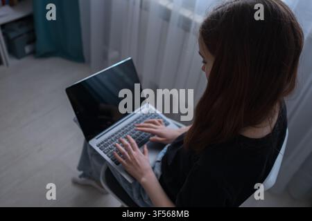 Young woman sitting with a laptop, focusing on the screen and typing on the keyboard, reflecting modern remote work, online learning, and digital Stock Photo