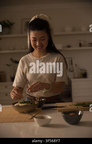 Adult Asian woman preparing meal in home kitchen, adding chopped herbs on wooden cutting board Stock Photo