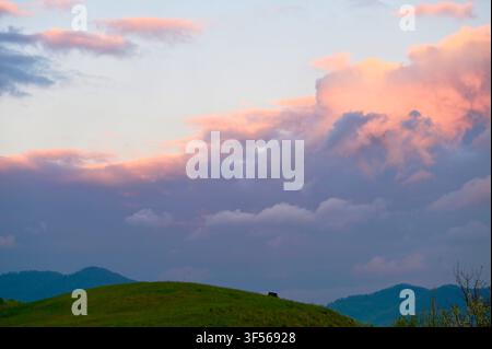 A scenic landscape featuring rolling green hills below a dramatic, colorful sky filled with glowing pink and purple sunset clouds during the evening twilight. Stock Photo