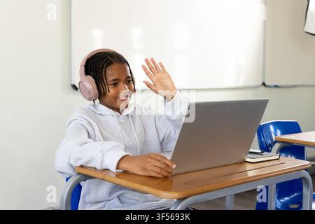 African American teen sitting in class at desk waving at silver laptop with pink headset Stock Photo
