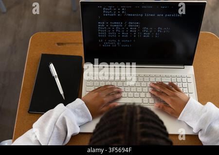 African American teen male typing code on silver laptop at wooden student desk with notebook pen Stock Photo