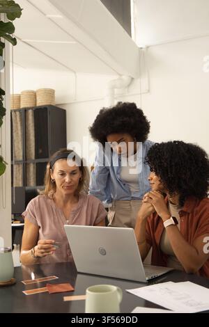 Diverse female coworkers collaborating at studio around laptop, reviewing color swatches and plans Stock Photo
