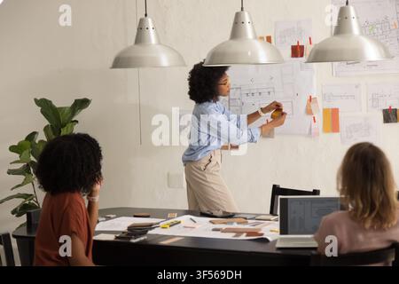 Diverse female coworkers pinning plans, reviewing material swatches, using laptop at studio table Stock Photo