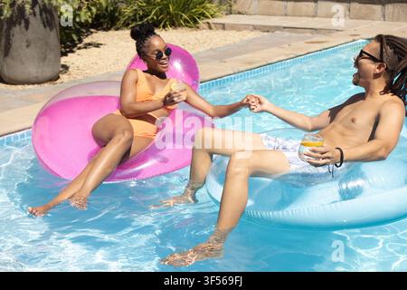 African American couple reclining on heart and ring floats in home pool, holding drinks and hands Stock Photo