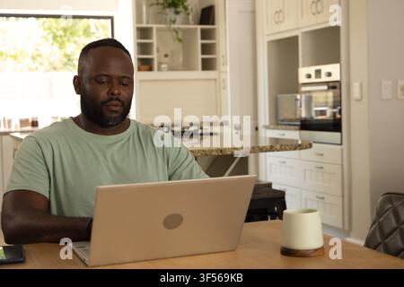 Mid adult African American man sitting at wooden dining table in modern kitchen using silver laptop Stock Photo