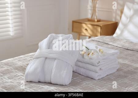 Clean bathrobe, towels and flowers on bed indoors Stock Photo