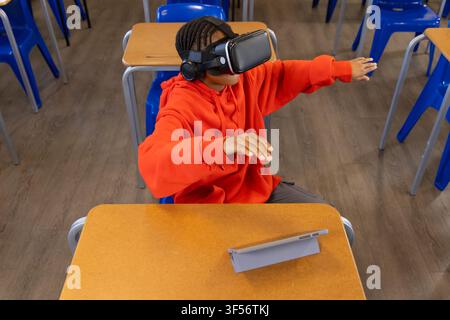 African American teen in orange hoodie using VR headset and headphones at school desk with tablet Stock Photo