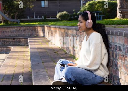 Girl child sitting on brick wall in sunny park in jeans headphones holding tablet-phone, copy space Stock Photo