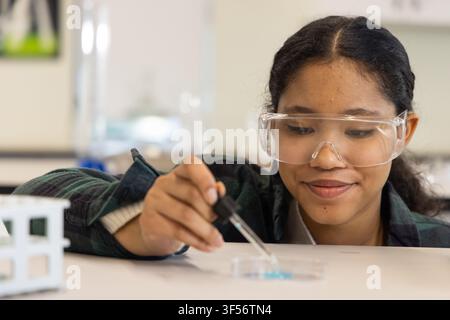 Female child in flannel and goggles using dropper above petri dish with blue-liquid at science lab Stock Photo